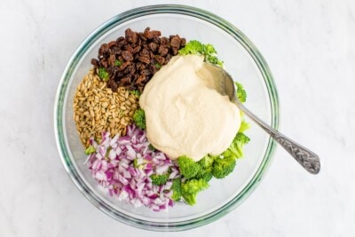 Ingredients for salad in a bowl: raw broccoli, raisins, sunflower seeds, red onion and creamy cashew dressing.