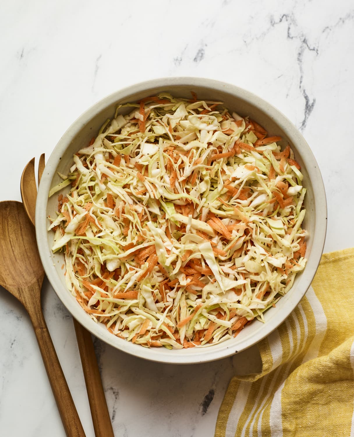 Serving bowl of apple cider vinegar slaw. Wood serving spoons and a dish cloth are next to the bowl.