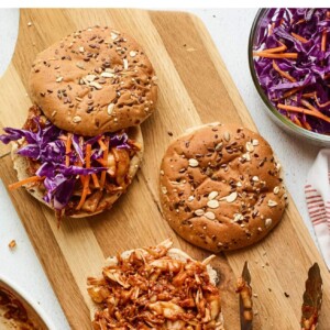 Wood cutting board with bbq jackfruit sandwiches being assembled.