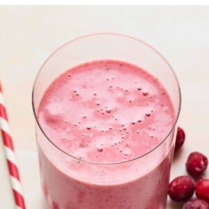 Glass with cranberry smoothie. A white and red paper straw and frozen cranberries are next to the glass.