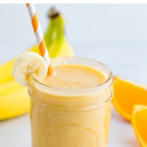 Orange banana smoothie in a glass jar, garnished with a banana slice and served with an orange and white striped paper straw.