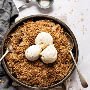 Round dish with an apple crisp topped with three scoops of vanilla ice cream. Two serving spoons are in the crisp.
