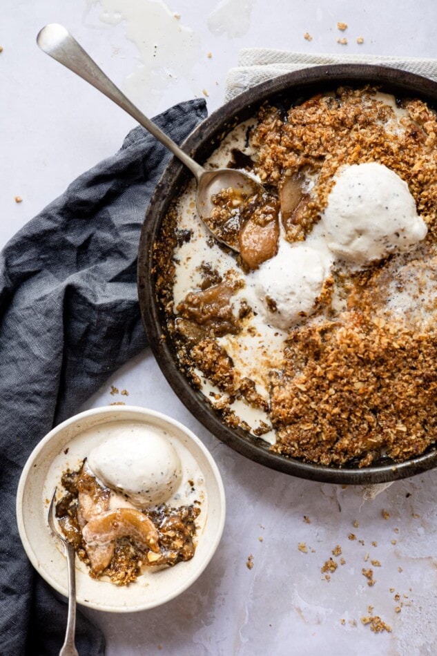 Apple crisp in a dish with a serving spoon and topped with ice cream. A bowl of apple crisp, ice cream and spoon is next to the large apple crisp.