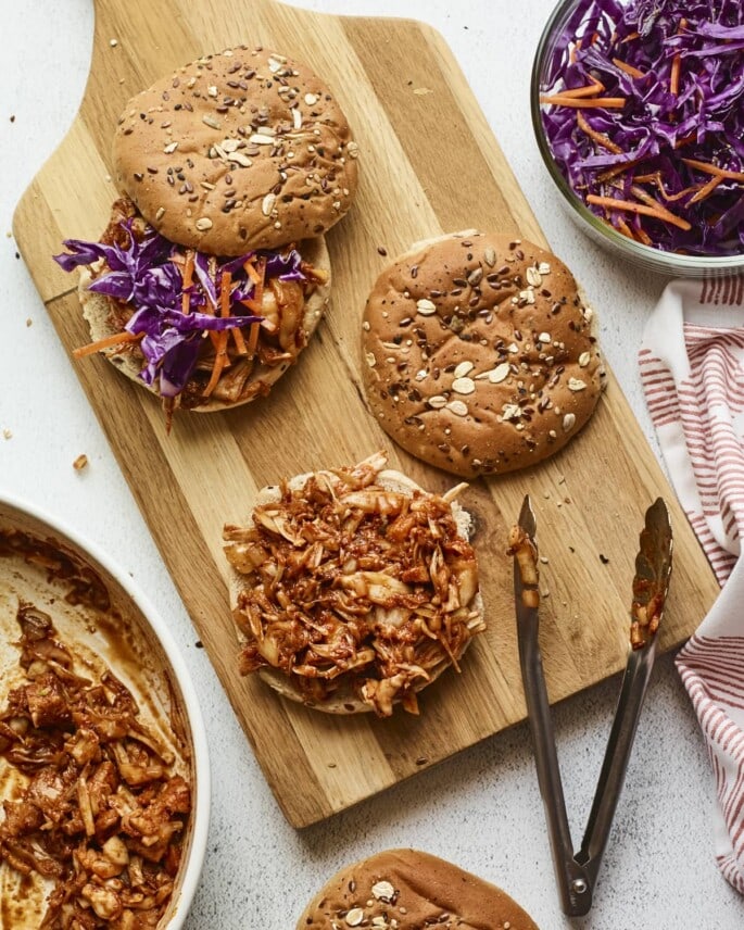 Wood cutting board with bbq jackfruit sandwiches being assembled.