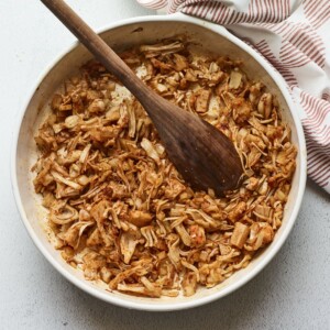 Bowl of bbq jackfruit. A wood spoon is in the bowl and dish towel is beside the bowl.