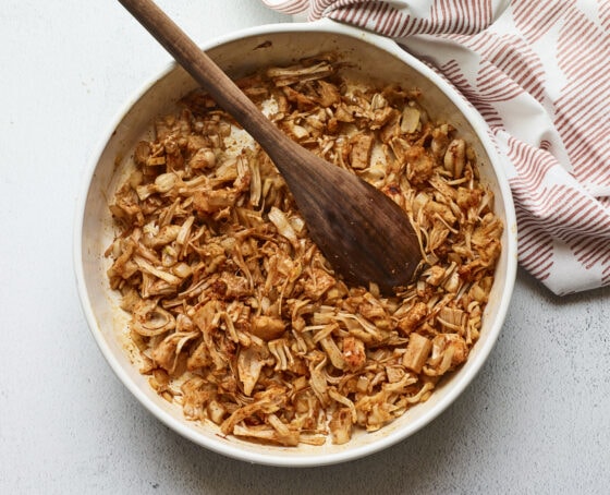 Bowl of bbq jackfruit. A wood spoon is in the bowl and dish towel is beside the bowl.