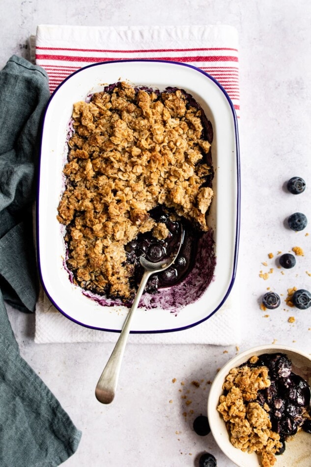 Blueberry crumble in a baking dish. A serving spoon as taken out a serving and put it in a bowl.