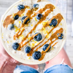 Woman holding a bowl of blueberry lemon overnight oats with a almond butter drizzle and gold spoon.