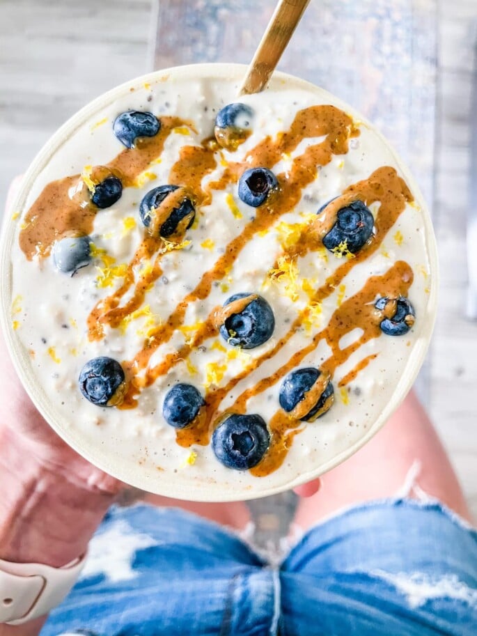Woman holding a bowl of blueberry lemon overnight oats with a almond butter drizzle and gold spoon. 