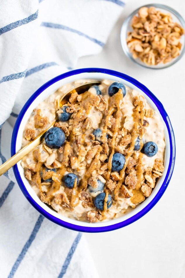 Blueberry overnight oats in a white bowl with blue rim. Topped with blueberries, crumble and nut butter.