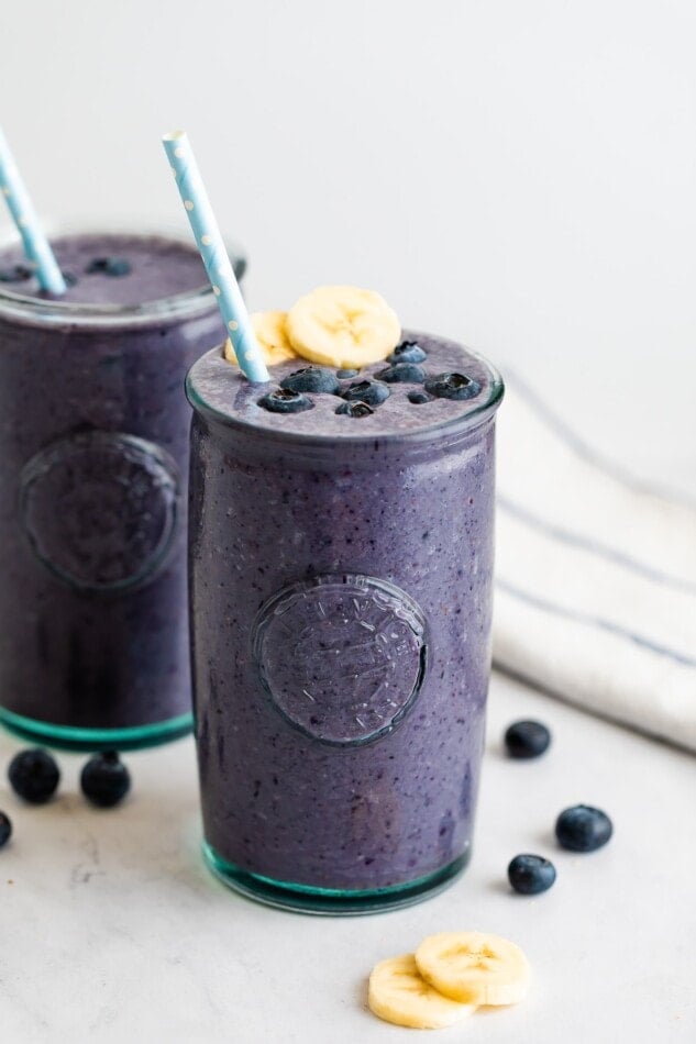 Blueberry smoothie in a glass topped with blueberries, banana slices and a straw.