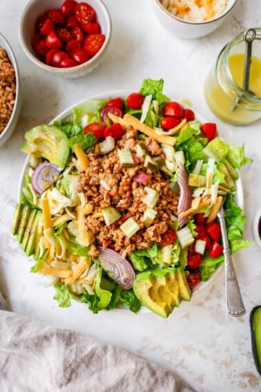 Cheeseburger salad topped with ground turkey, avocado, cheddar, tomatoes and onions. A bowl of ground turkey, tomatoes, cheddar cheese and honey mustard dressing are beside the bowl.
