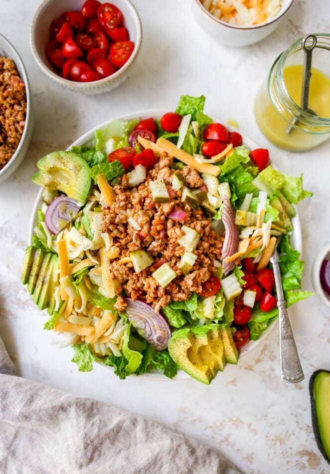 Cheeseburger salad topped with ground turkey, avocado, cheddar, tomatoes and onions. A bowl of ground turkey, tomatoes, cheddar cheese and honey mustard dressing are beside the bowl.