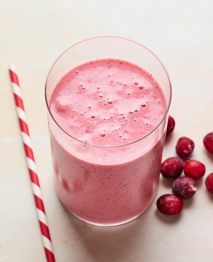 Glass with cranberry smoothie. A white and red paper straw and frozen cranberries are next to the glass.