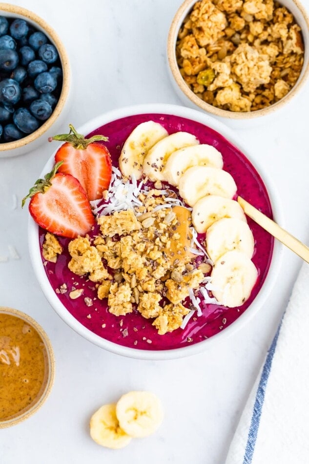 Dragon fruit smoothie bowl topped with granola, banana slices and a strawberry with a gold spoon.