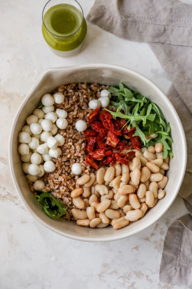 Mozzarella balls, farro, arugula, sun-dried tomatoes and beans in a bowl. A jar of basil vinaigrette is to the side of the bowl.