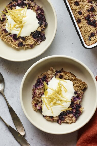 Servings of blueberry lemon baked oatmeal in two bowls and topped with yogurt and lemon zest.