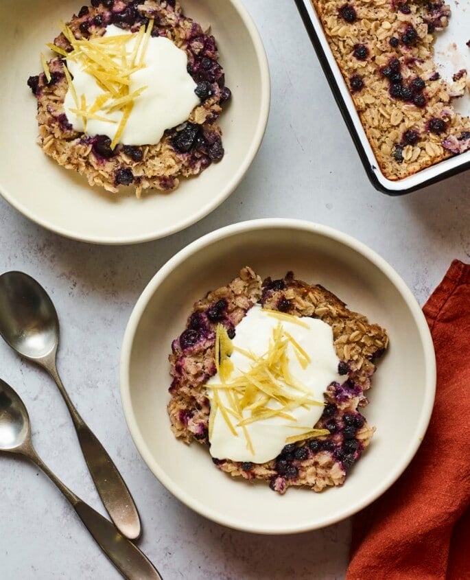 Servings of blueberry lemon baked oatmeal in two bowls and topped with yogurt and lemon zest.