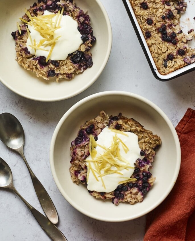 Servings of blueberry lemon baked oatmeal in two bowls and topped with yogurt and lemon zest.