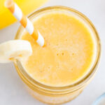 Orange banana smoothie in a glass jar, garnished with a banana slice and served with an orange and white striped paper straw.