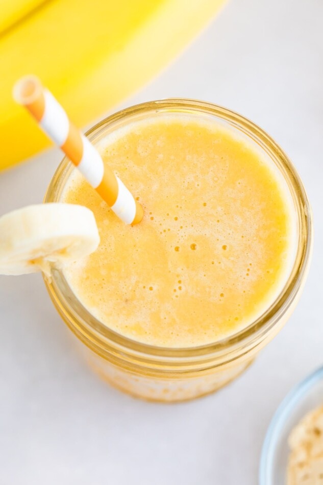 Orange banana smoothie in a glass jar, garnished with a banana slice and served with an orange and white striped paper straw.