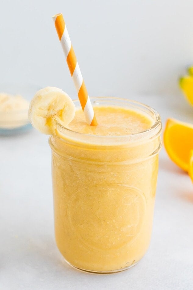 Orange banana smoothie in a glass jar, garnished with a banana slice and served with an orange and white striped paper straw.
