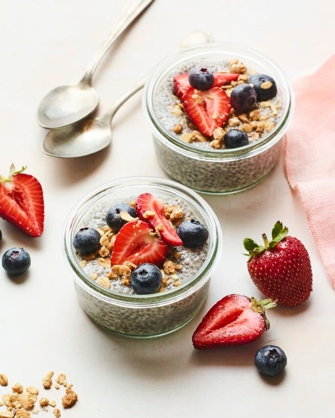 Two glass jars with chia pudding topped with fruit and granola. Strawberries, blueberries and spoons are on the table around the jars.