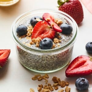 Jar of chia pudding topped with berries and granola. More granola and berries are scattered on the table around the jar.