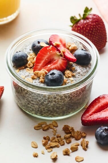 Jar of chia pudding topped with berries and granola. More granola and berries are scattered on the table around the jar.