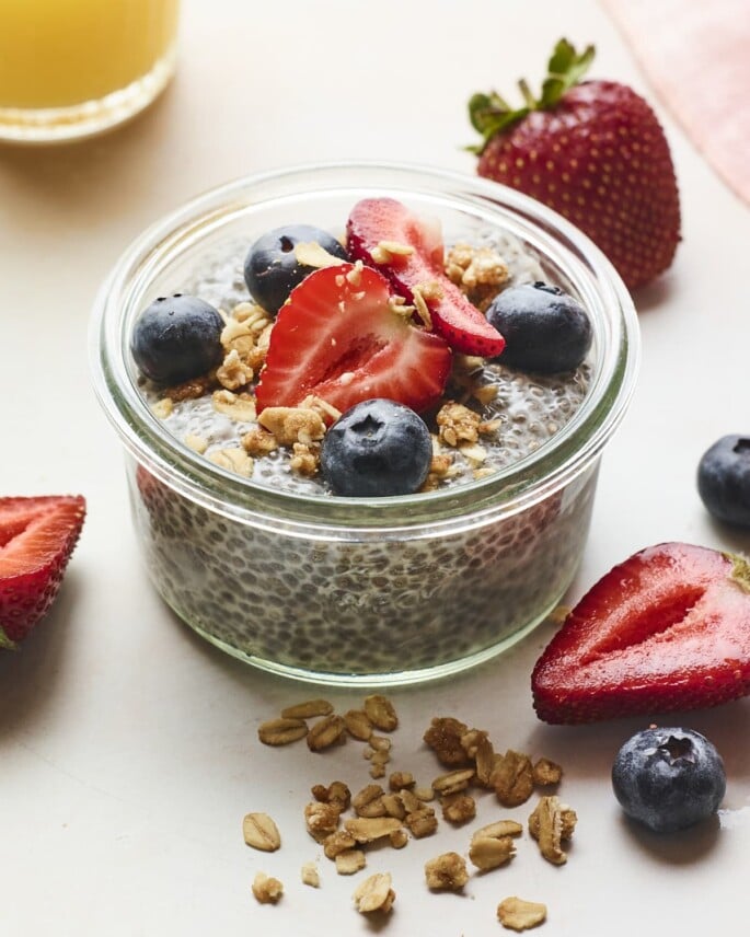 Jar of chia pudding topped with berries and granola. More granola and berries are scattered on the table around the jar.