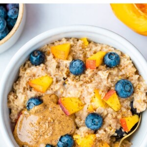 Blueberry peach oatmeal in bowl with blueberries and fresh chopped peaches on top with a little almond butter on top as well.