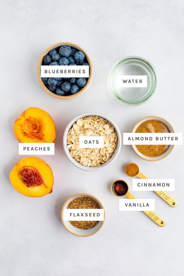 Ingredients measured out to make blueberry peach oatmeal: blueberries, water, peaches, oats, almond butter, cinnamon, vanilla and flaxseed.