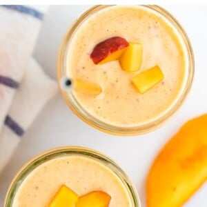 Two mason jars with peach smoothies. Smoothies are garnished with diced peaches and gold straws.