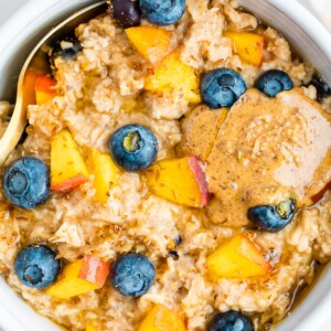 Overhead close up of blueberry peach oatmeal in a white bowl with blueberries and fresh chopped peaches on top with a little almond butter on top as well.