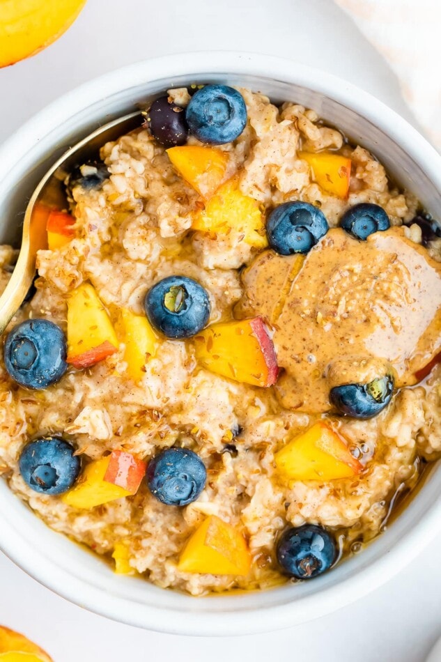 Overhead close up of blueberry peach oatmeal in a white bowl with blueberries and fresh chopped peaches on top with a little almond butter on top as well.