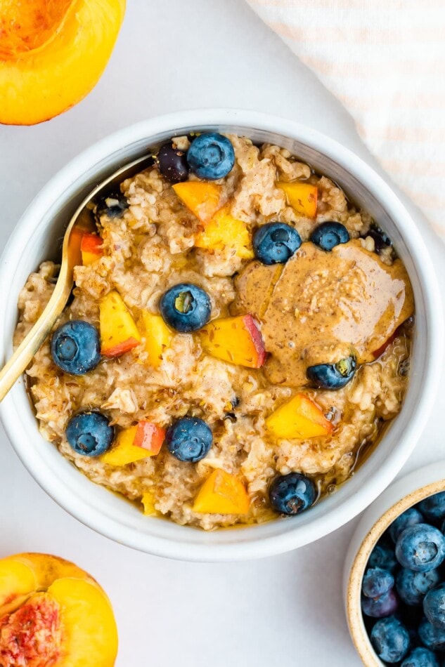 Overhead shot of blueberry peach oatmeal in a white bowl with blueberries and fresh chopped peaches on top with a little almond butter on top as well.