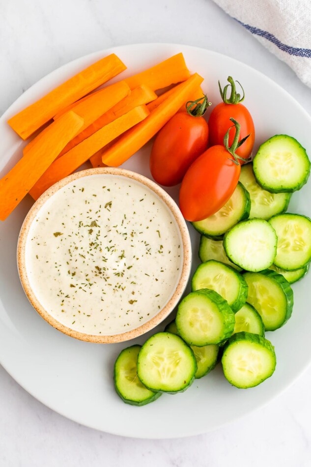 Vegan ranch in a small bowl with fresh veggies around it for dipping. All on a white plate. 