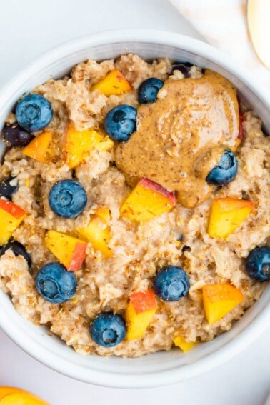 Blueberry peach oatmeal in bowl with blueberries and fresh chopped peaches on top with a little almond butter on top as well.