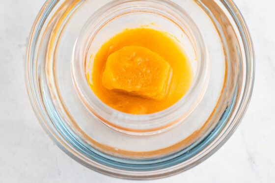 A frozen peach puree cube in a small glass container, being submerged in a water bath to defrost.