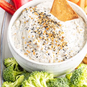 Plate with veggies and pita chips. On the plate is a bowl of everything bagel dip and a pita chip is in the dip.