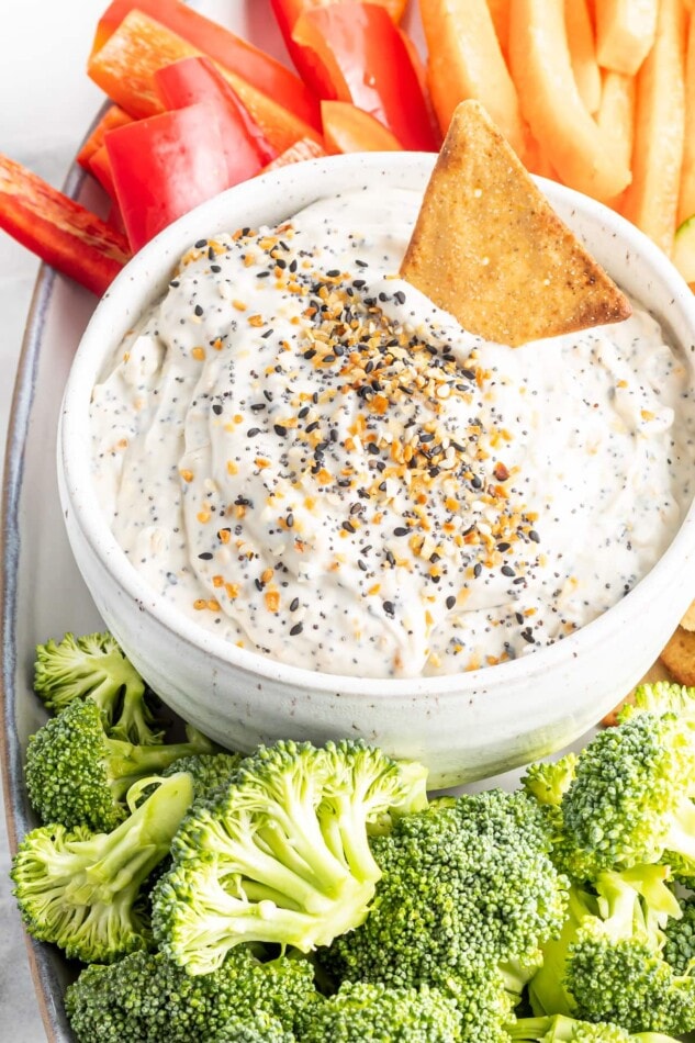 Plate with veggies and pita chips. On the plate is a bowl of everything bagel dip and a pita chip is in the dip.