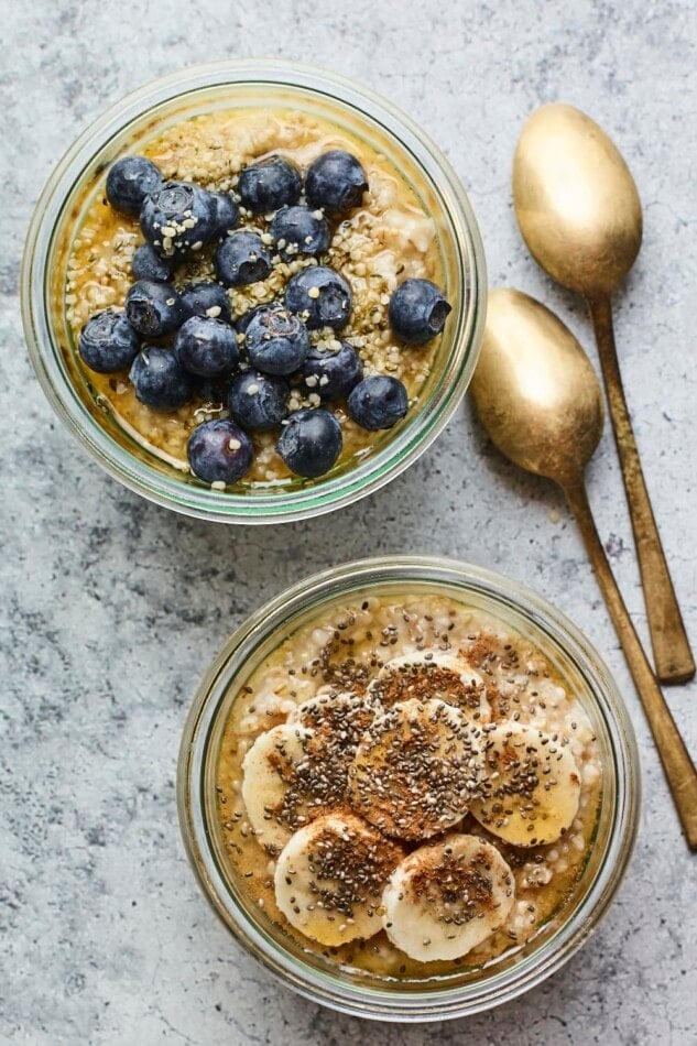 Two glass bowls of overnight steel cut oats. One it topped with blueberries and the other is topped with banana, maple and chia seeds.