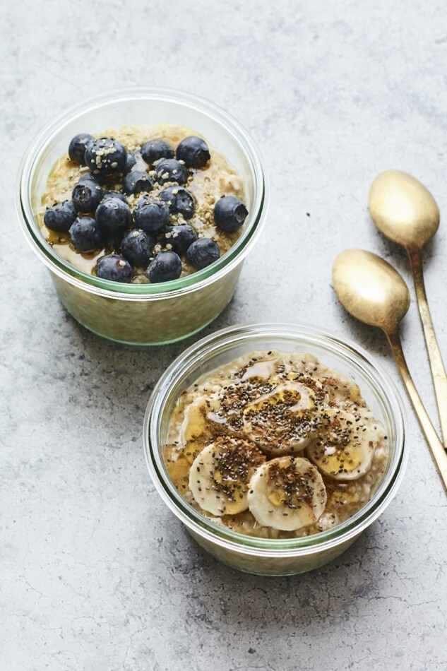 Two glass bowls of overnight steel cut oats. One it topped with blueberries and the other is topped with banana, maple and chia seeds.