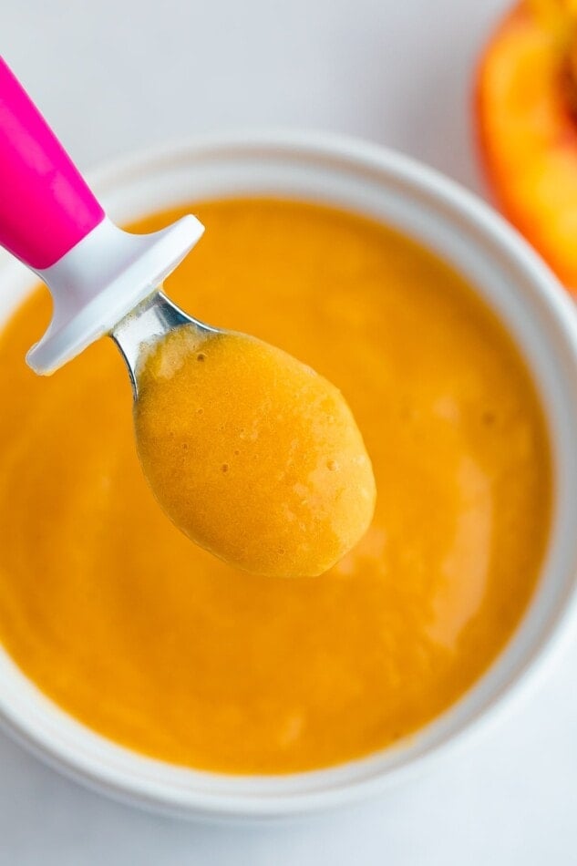 Up close shot of peach puree on a pink baby spoon hovering over a white bowl with peach baby food.