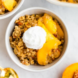 A white bowl with peach crisp portion and a dollop of coconut whipped cream on top.