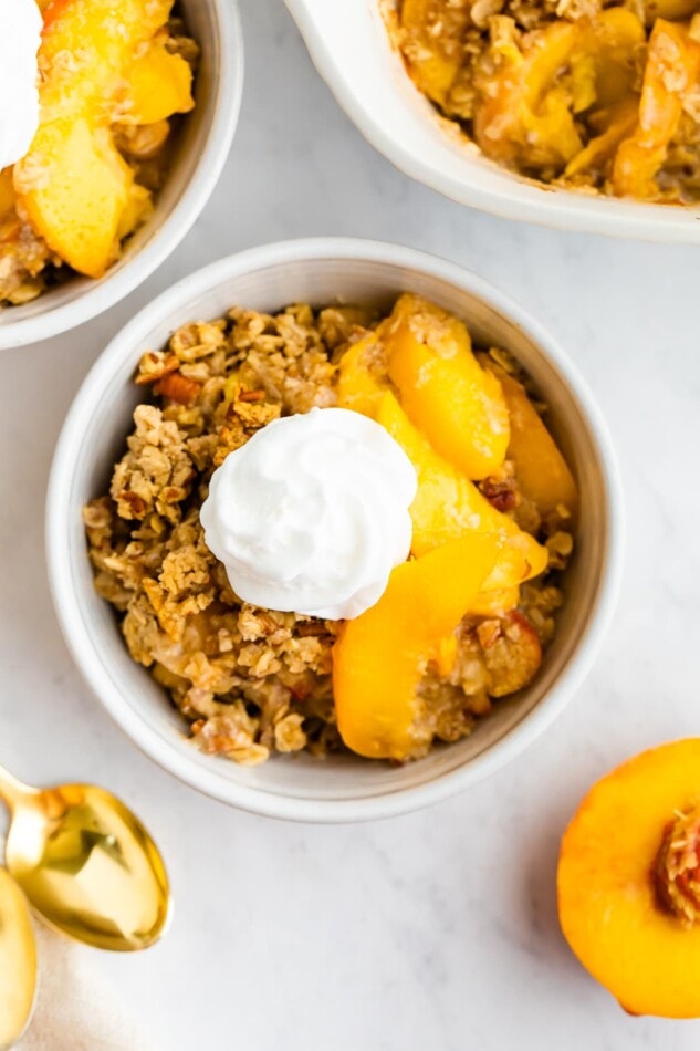 A white bowl with peach crisp portion and a dollop of coconut whipped cream on top.