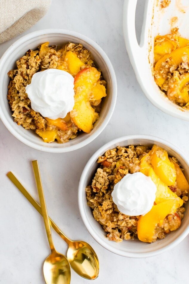 Two bowls of peach crisp, each in white bowls with a dollop of whipped cream on top and two gold spoons on the side.