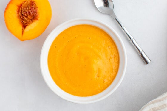 Pureed peaches in a white bowl on a marble surface with a small silver spoon on the side.