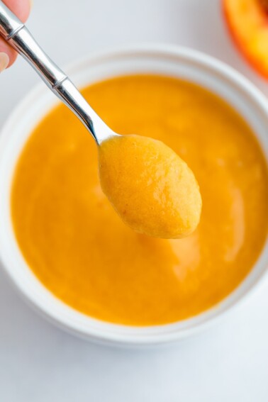 Up close shot of peach puree on a small sliver spoon hovering over a white bowl with peach puree.