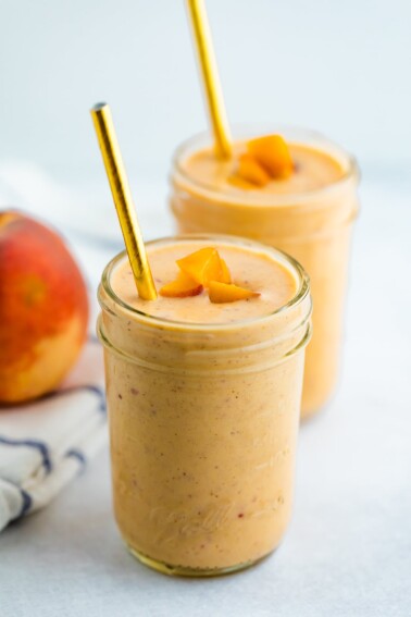 Two glass mason jars of peach smoothies. Two gold straws are in the smoothie and the smoothies are topped with chopped peached.